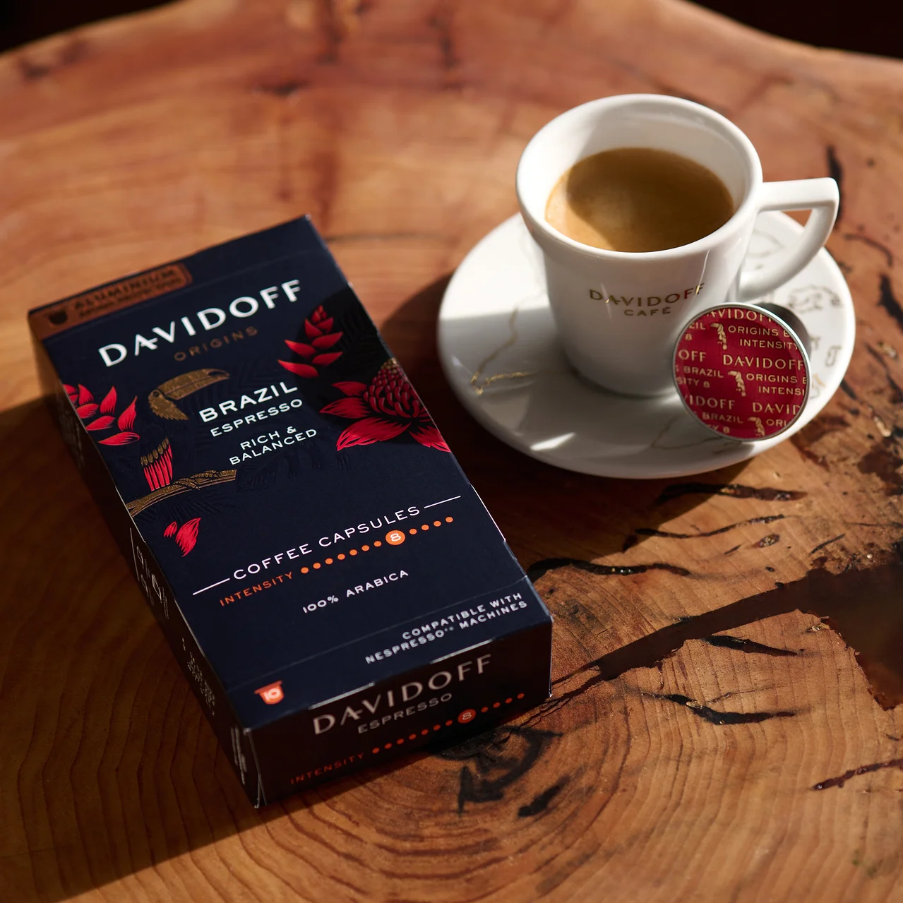Ahşap bir masanın üzerinde bir paket DAVIDOFF CAFÉ ORIGINS Brazil Espresso, bir fincan espresso ve bir kapsül var.