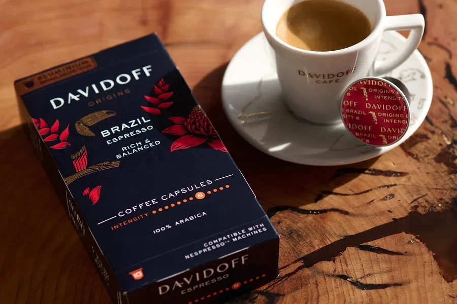 Ahşap bir masanın üzerinde bir paket DAVIDOFF CAFÉ ORIGINS Brazil Espresso, bir fincan espresso ve bir kapsül var.