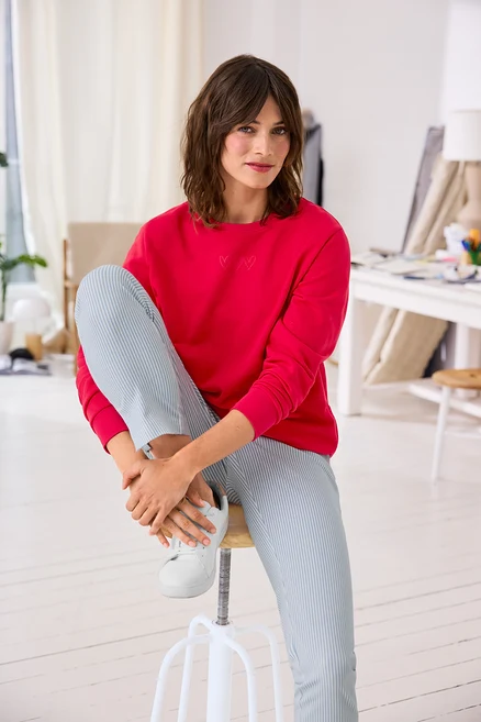 Kahverengi saçlı bir kadın beyaz bir taburede oturuyor, pembe kalp işlemeli bir sweatshirt, açık mavi çizgili pantolon ve beyaz spor ayakkabı giymiş, ayağını tutuyor.