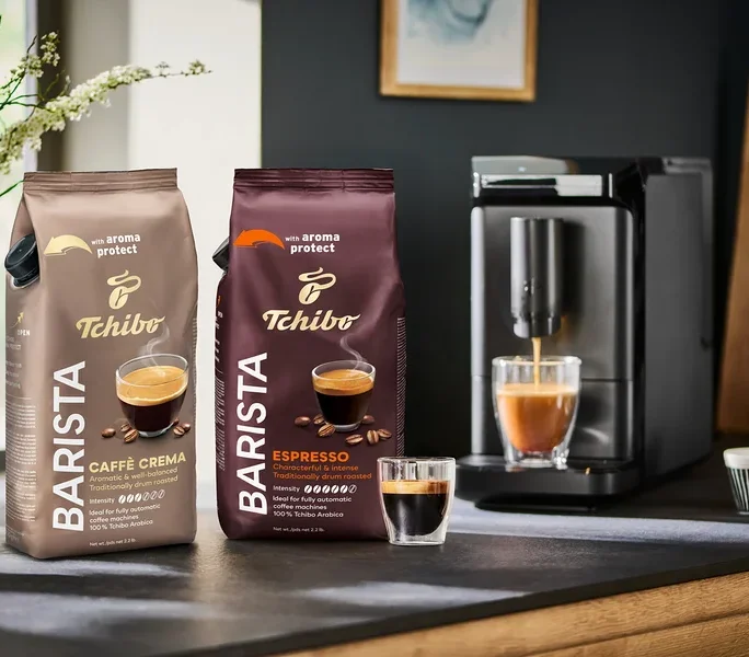 Fincanların ve kahve makinesinin yanında iki paket Tchibo Barista Caffe Crema ve Espresso kahvesi. Siyah »Esperto mini« kahve makinesi de görülebilir.