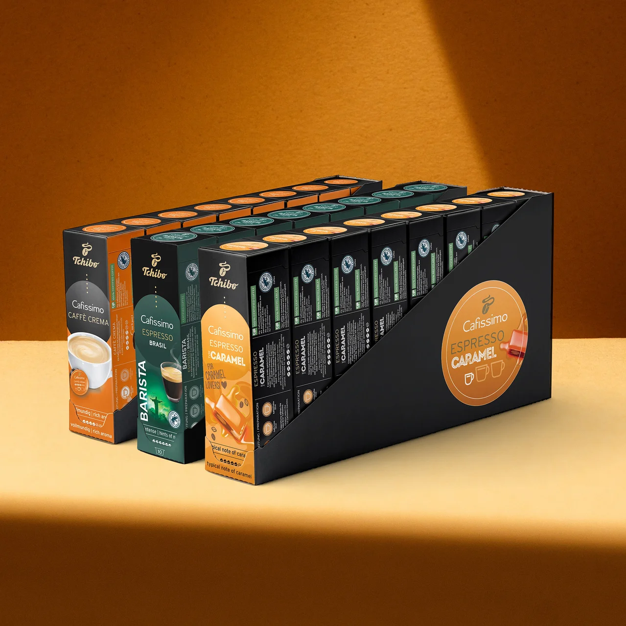 Siyah bir standda üç sıra halinde dizilmiş Tchibo Cafissimo kahve kapsülü paketleri: Caffè Crema, Espresso Brasil Barista ve Espresso Caramel.
