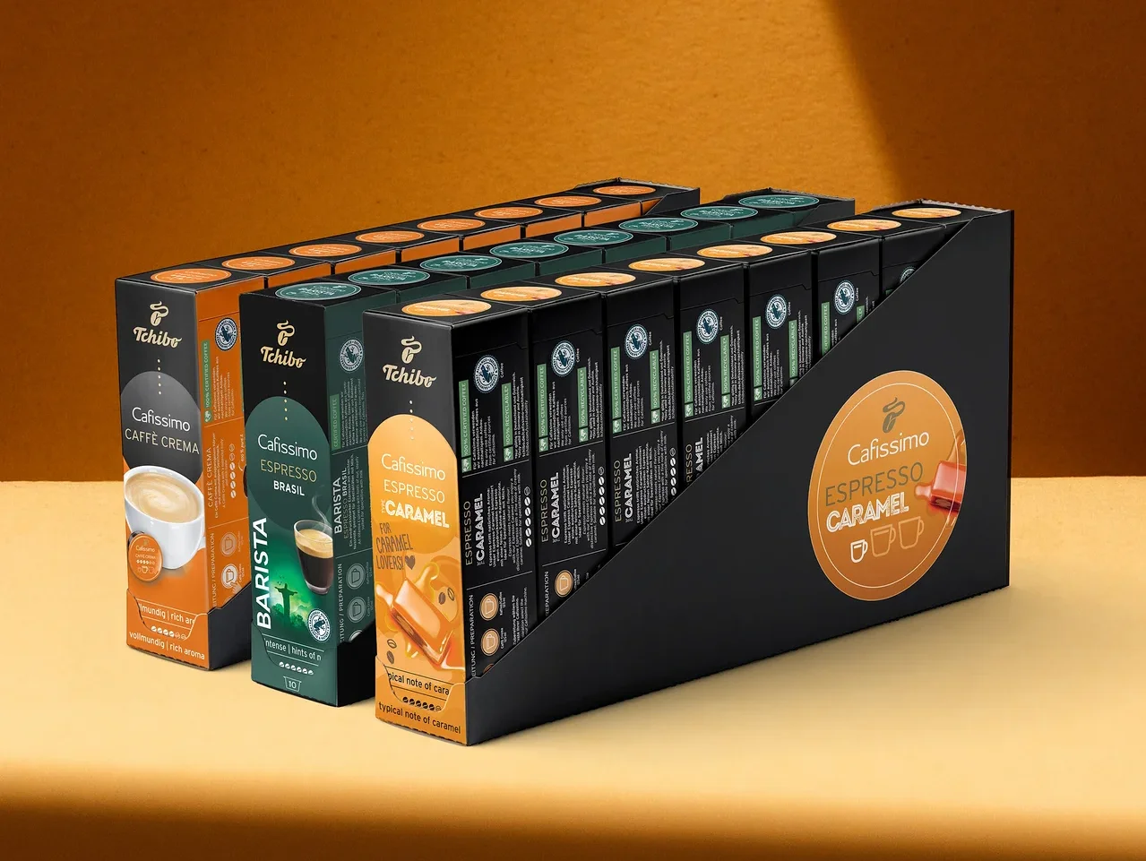 Siyah bir standda üç sıra halinde dizilmiş Tchibo Cafissimo kahve kapsülü paketleri: Caffè Crema, Espresso Brasil Barista ve Espresso Caramel.