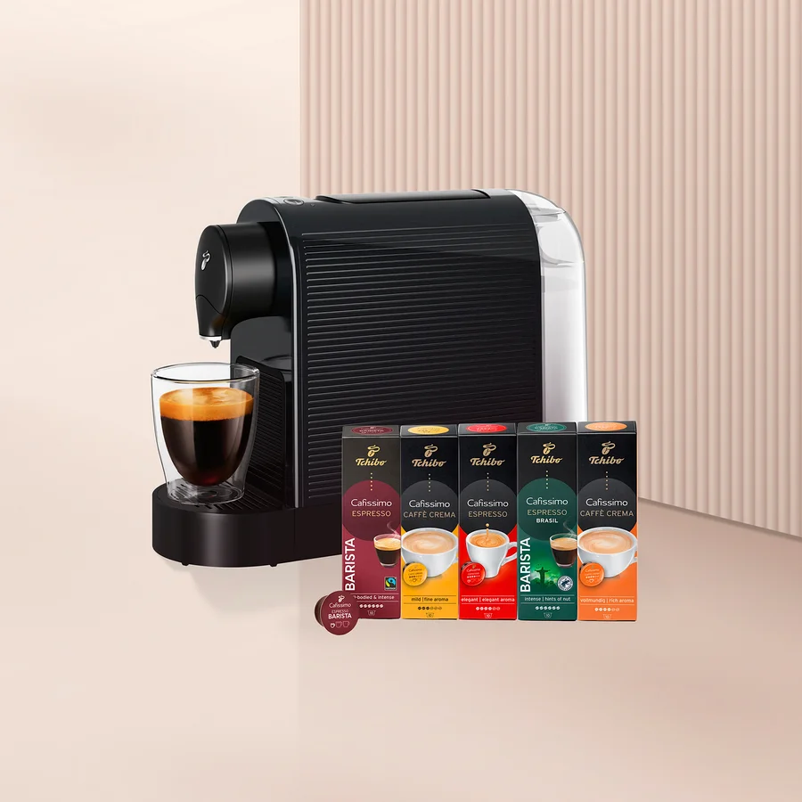 Siyah beyaz kapsül kahve makinesi, altında cam bir fincan kahve ve yanında beş kutu Tchibo Cafissimo kahve kapsülü ile bir adet Barista kapsülü.