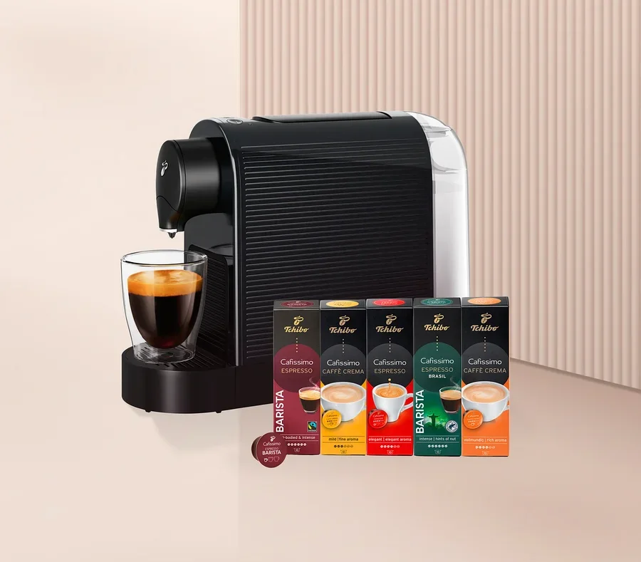 Siyah beyaz kapsül kahve makinesi, altında cam bir fincan kahve ve yanında beş kutu Tchibo Cafissimo kahve kapsülü ile bir adet Barista kapsülü.