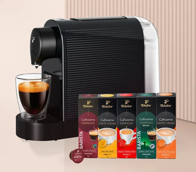Siyah beyaz kapsül kahve makinesi, altında cam bir fincan kahve ve yanında beş kutu Tchibo Cafissimo kahve kapsülü ile bir adet Barista kapsülü.