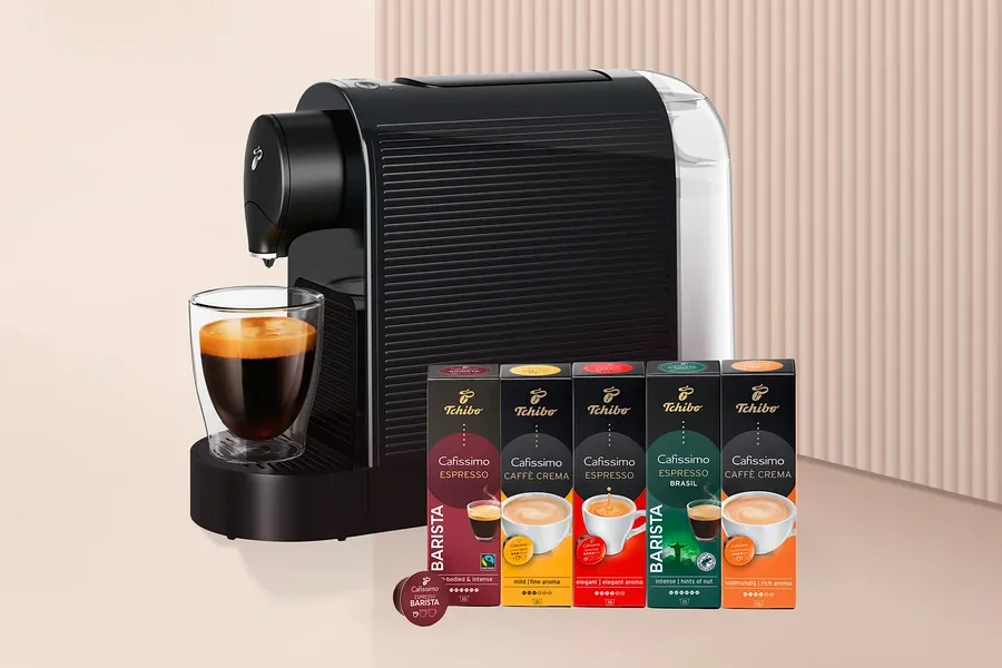 Siyah beyaz kapsül kahve makinesi, altında cam bir fincan kahve ve yanında beş kutu Tchibo Cafissimo kahve kapsülü ile bir adet Barista kapsülü.