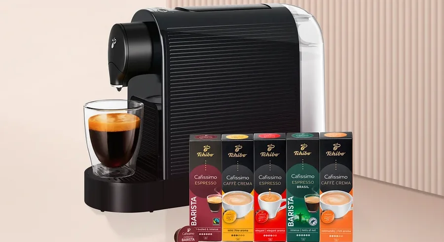 Siyah beyaz kapsül kahve makinesi, altında cam bir fincan kahve ve yanında beş kutu Tchibo Cafissimo kahve kapsülü ile bir adet Barista kapsülü.