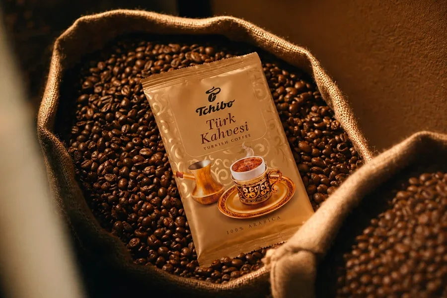 Cezve ve fincan illüstrasyonlu altın rengi Tchibo Türk Kahvesi paketi, jüt çuvaldaki kahve çekirdeklerinin üzerinde duruyor. „%100 ARABICA“ yazısı görünüyor.