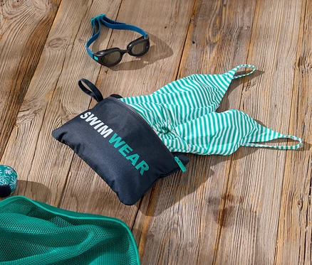 Ahşap bir yüzey üzerinde, üzerinde "Swimwear" yazan bir çanta, yeşil ve beyaz çizgili bir mayo, bir çift yüzme gözlüğü ve yeşil bir file bulunan yüzme aksesuarlarının çekimi.