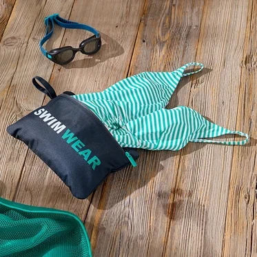 Ahşap bir yüzey üzerinde, üzerinde "Swimwear" yazan bir çanta, yeşil ve beyaz çizgili bir mayo, bir çift yüzme gözlüğü ve yeşil bir file bulunan yüzme aksesuarlarının çekimi.