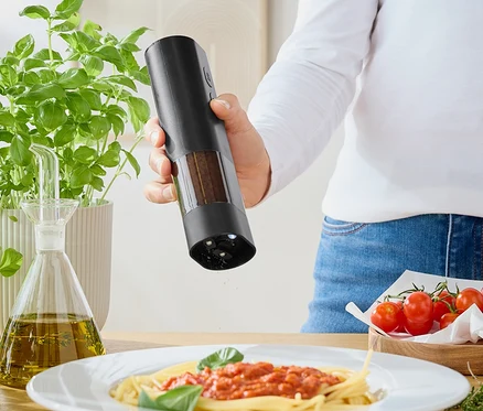 Kadın, değirmenden domates soslu spagettinin üzerine baharat serpiyor. Masanın üzerinde fesleğen, zeytinyağı ve domatesler var.