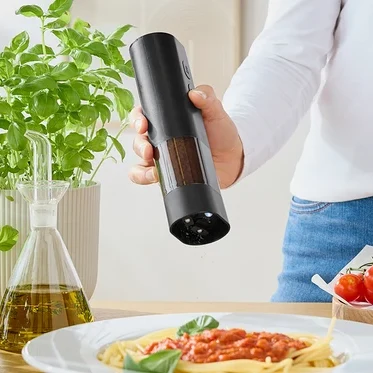 Kadın, değirmenden domates soslu spagettinin üzerine baharat serpiyor. Masanın üzerinde fesleğen, zeytinyağı ve domatesler var.