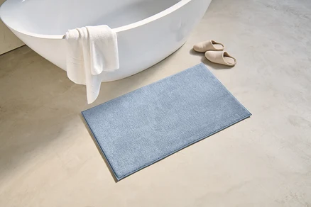 Beyaz havlunun asılı olduğu küvetin yanında, yerde 60 x 100 cm boyutlarında mavi bir banyo paspası ve bej terlikler bulunmaktadır.
