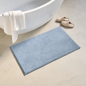 Beyaz havlunun asılı olduğu küvetin yanında, yerde 60 x 100 cm boyutlarında mavi bir banyo paspası ve bej terlikler bulunmaktadır.