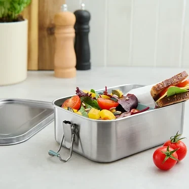 Paslanmaz çelik yemek kutusunda salata ve sandviç, yanında iki domates duruyor.