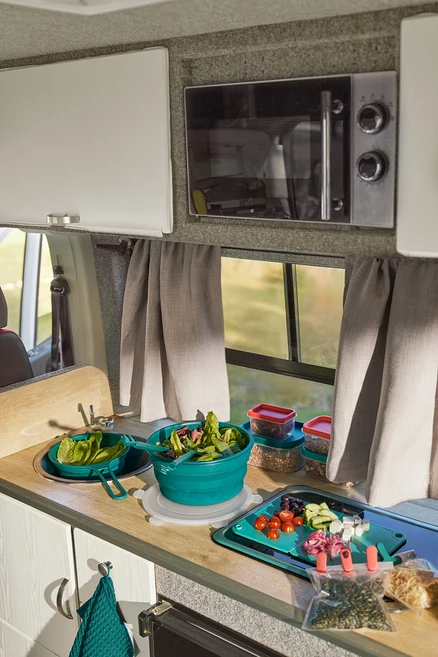 Karavan mutfak tezgahında lavaboda yer tasarruflu süzgeç, salata ve 2'si 1 arada spatula-maşa içeren katlanabilir salata kasesi. Katlanabilir kesme tahtasında doğranmış sebzeler var. Yanında iki aroma klipsi ve iki saklama kabı. Pencerenin üzerinde bir mikrodalga fırın bulunuyor.