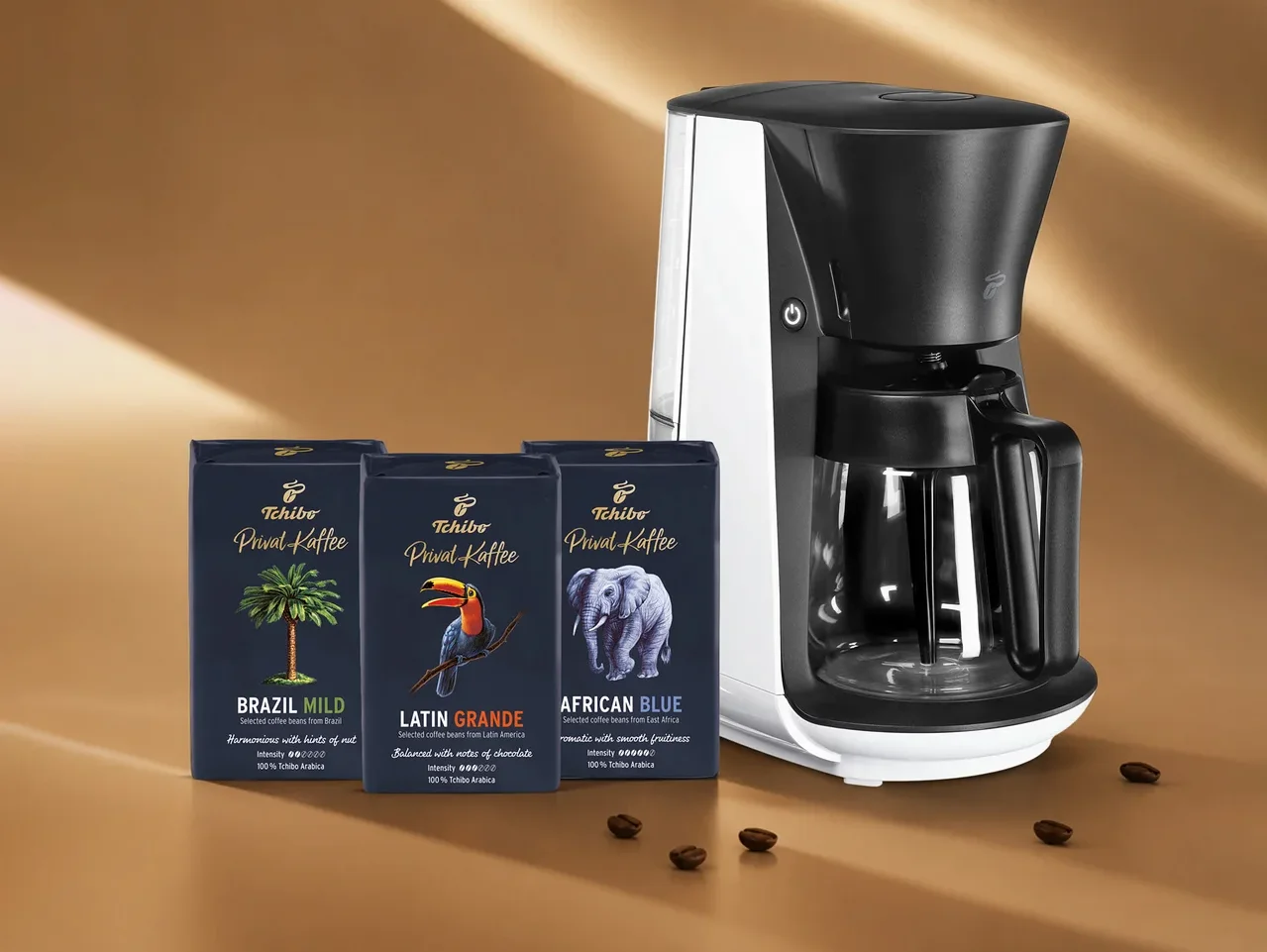 Beyaz ve siyah bir kahve makinesi, palmiye ağacı, tukan ve fil motifli üç koyu mavi Tchibo Privat Kaffee paketiyle birlikte kahverengi bir zeminde, etrafa saçılmış kahve çekirdekleriyle duruyor.