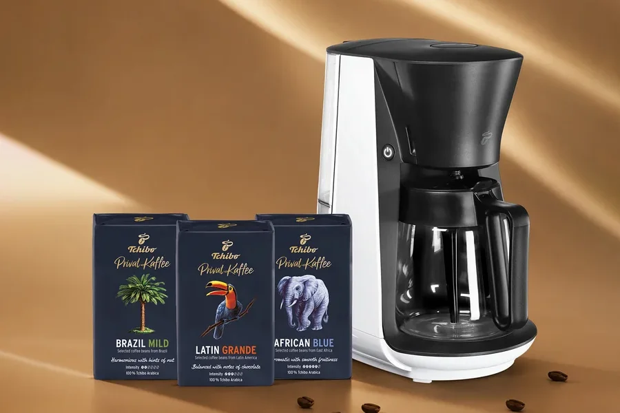 Beyaz ve siyah bir kahve makinesi, palmiye ağacı, tukan ve fil motifli üç koyu mavi Tchibo Privat Kaffee paketiyle birlikte kahverengi bir zeminde, etrafa saçılmış kahve çekirdekleriyle duruyor.