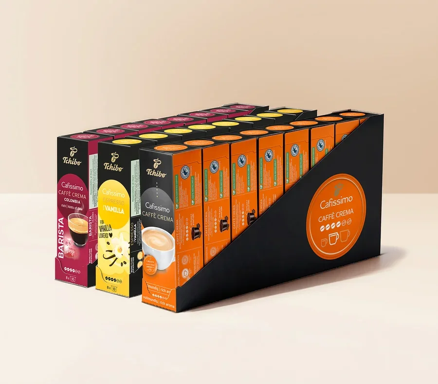 Siyah bir teşhir standında birden fazla sıra Tchibo Cafissimo kahve kapsülü kutusu bulunuyor. Görünen çeşitler arasında pembe Caffè Crema Colombia, sarı Espresso Vanilla ve turuncu Caffè Crema kutuları yer alıyor. Standın yanında turuncu bir Cafissimo logosu var.