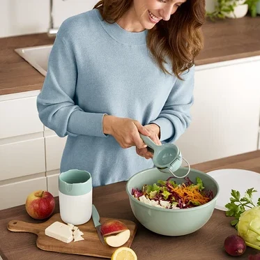 Kadın salata kasesine limon sıkıyor. Masada elma, peynir, badem ve sebzeler var.