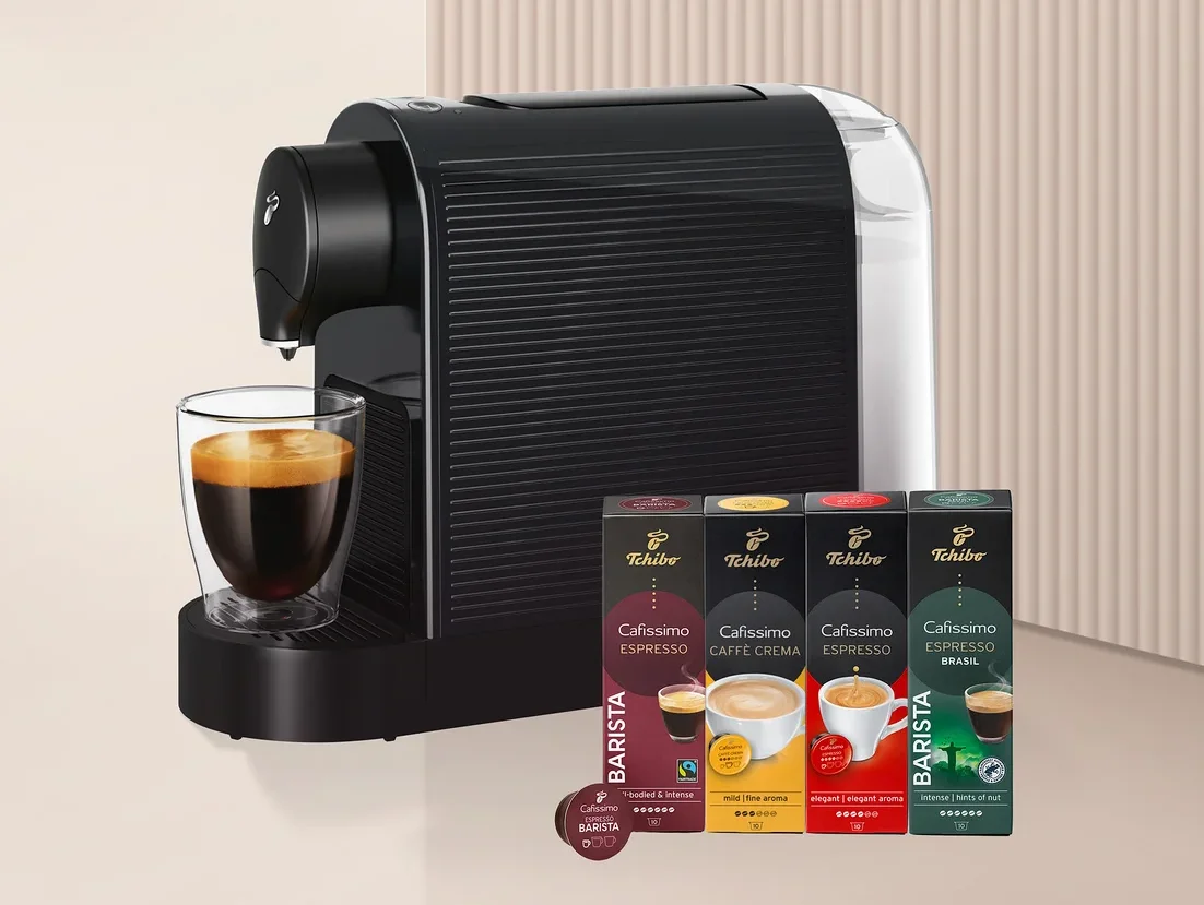 Siyah beyaz bir Tchibo Cafissimo kahve makinesi, şeffaf bir cam bardağa espresso dolduruyor. Yanında dört kutu Tchibo Cafissimo Barista Edition kapsülü (Espresso, Caffè Crema, Espresso, Espresso Brasil) ve bir adet tek kapsül bulunuyor.