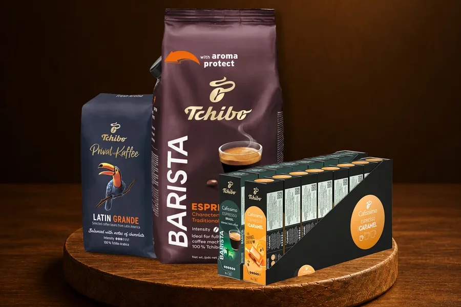 Ahşap tepside üç Tchibo kahve ürünü. Solda "Privat Kaffee Latin Grande" çekirdek kahve, arkada "Barista Espresso" çekirdek kahve, sağda "Cafissimo Espresso Caramel" kapsülleri.