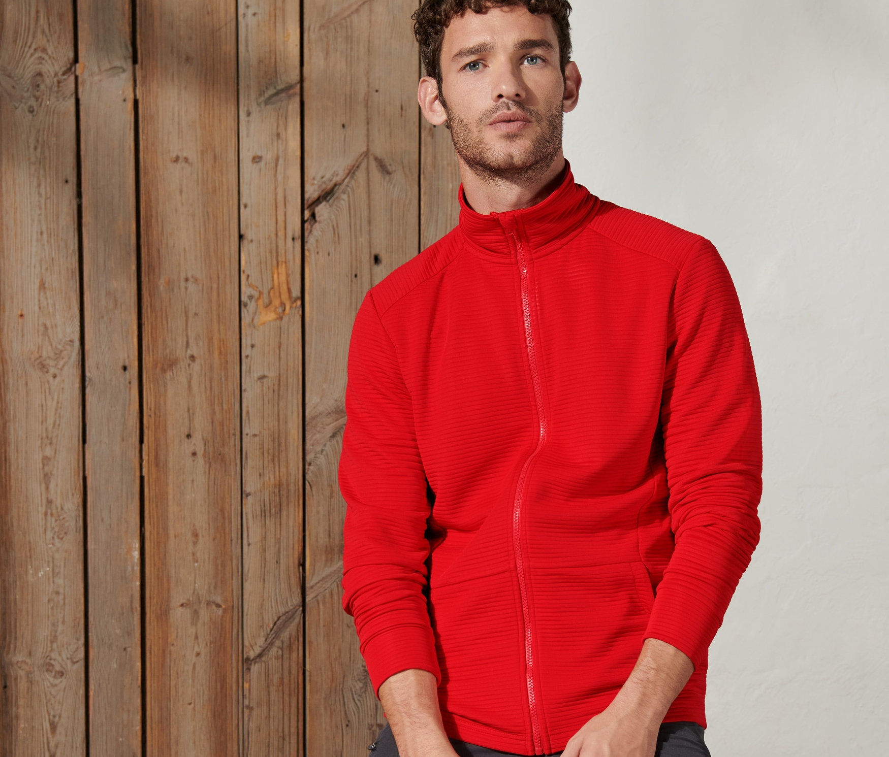 Adam ahşap bir kiriş üzerinde oturuyor ve koyu turuncu fonksiyonel bir ceket giyiyor.