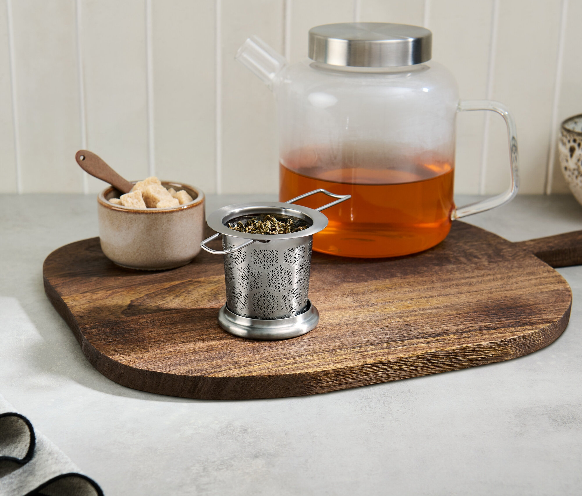 Çaylı çay süzgeci, çaylı cam çaydanlığın yanında ahşap bir tahtanın üzerinde duruyor.