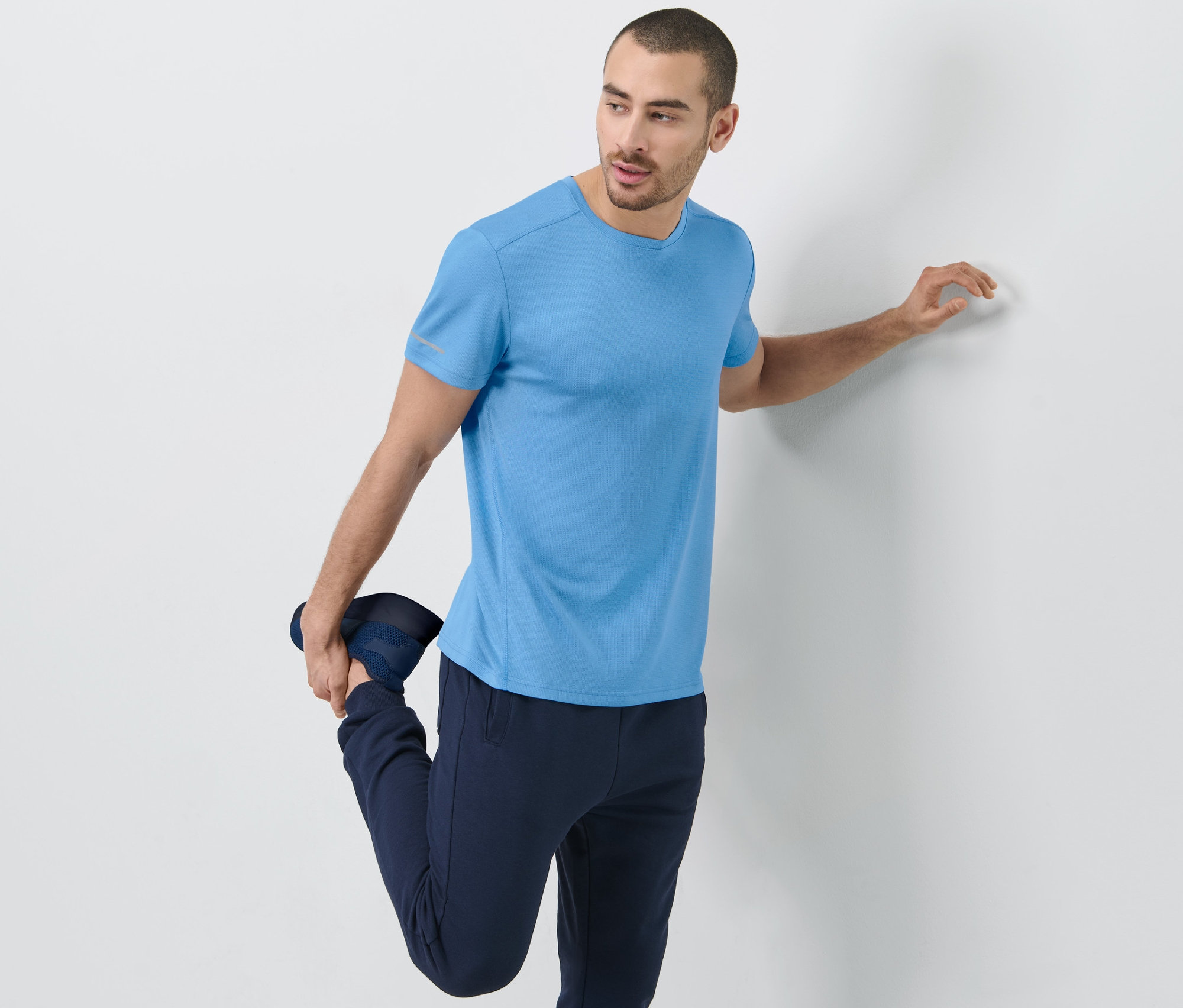 Adam bacağını esnetiyor, gök mavisi fonksiyonel tişört giyiyor.