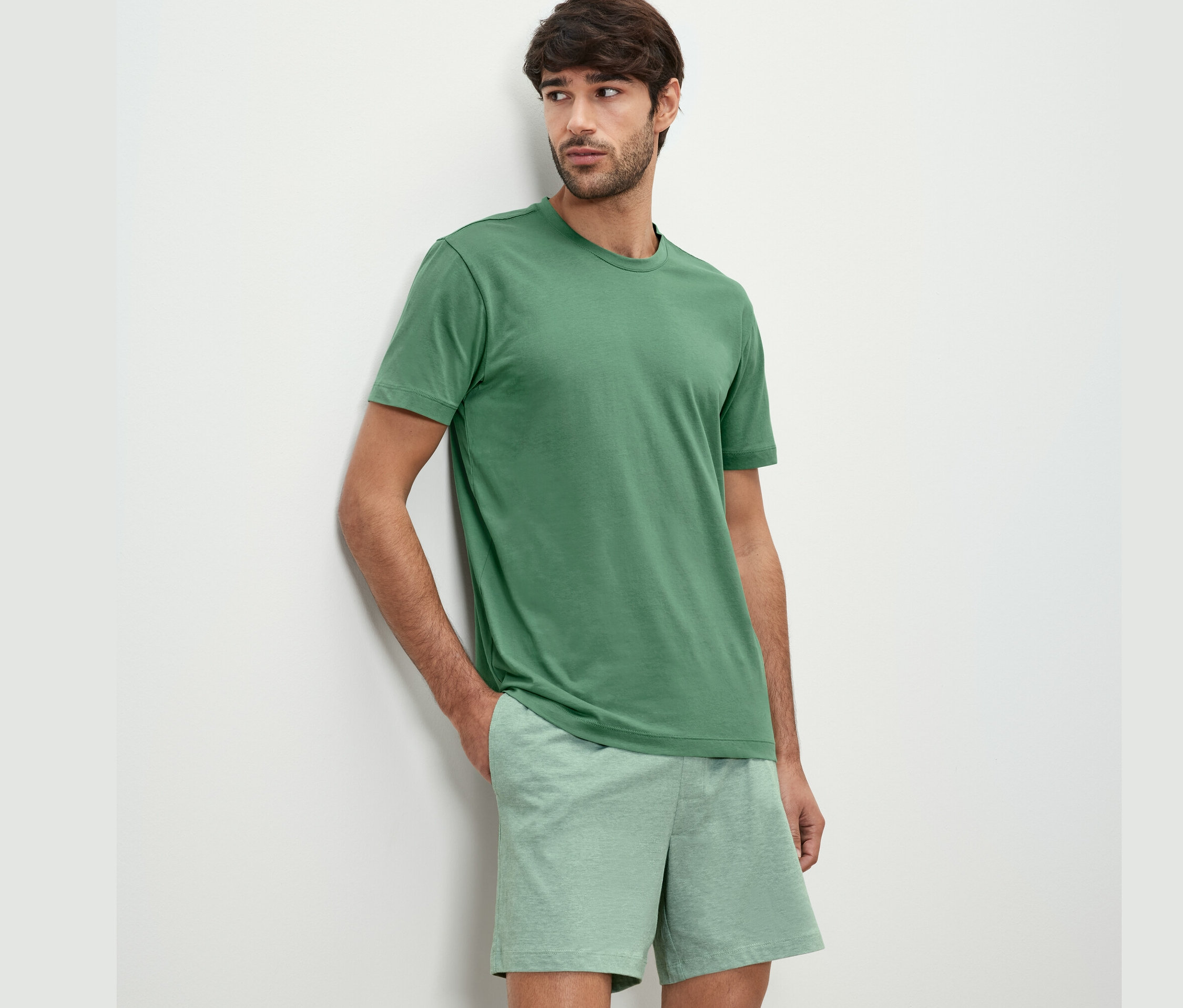 Duvara yaslanmış şortlu pijama takımı giyen bir adam.