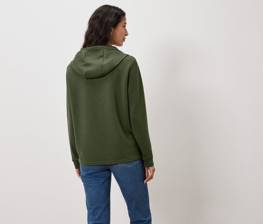 Kadın kameraya arkası dönük, yeşil bir kapüşonlu ve kot pantolon giyiyor.