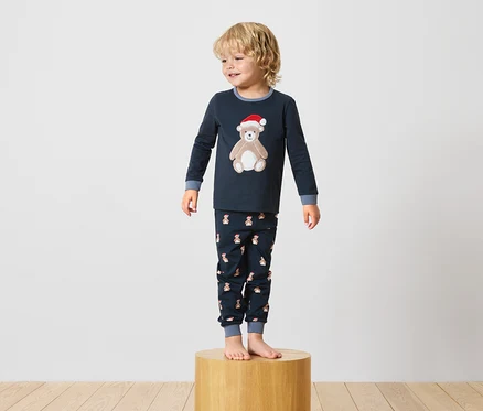 Ayakta duran bir çocuk, üzerinde oyuncak ayı deseni olan lacivert bir pijama giyiyor ve ahşap bir sehpanın üzerinde duruyor.