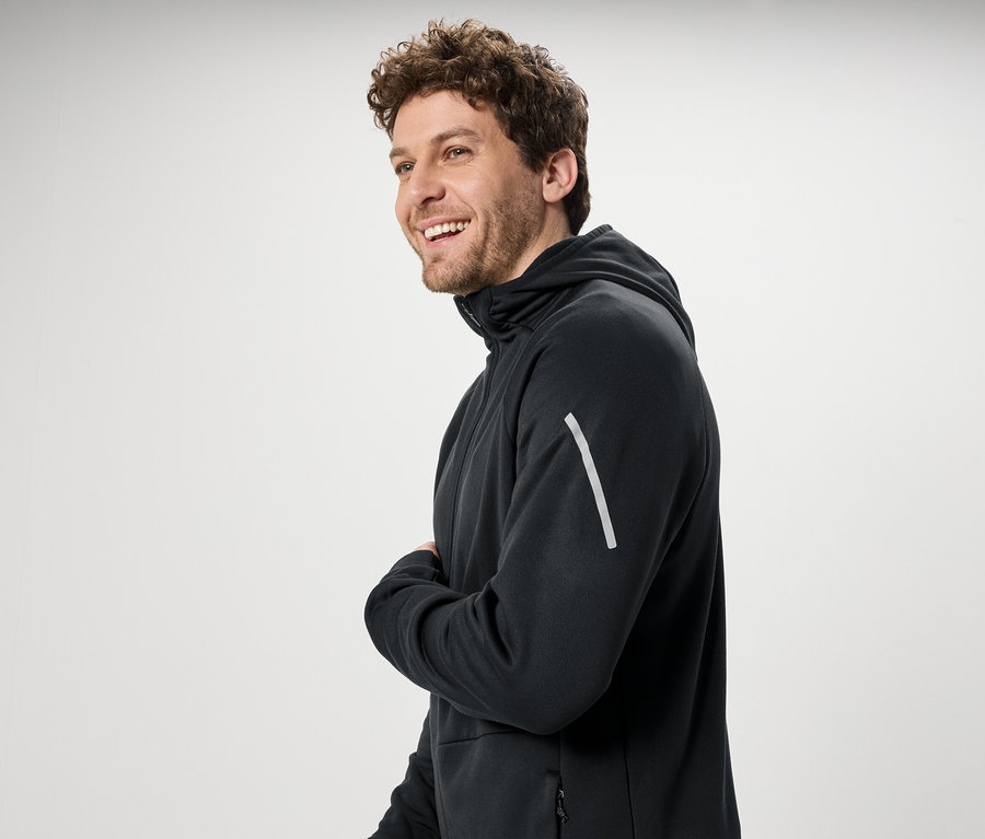 Kıvırcık saçlı gülümseyen bir adam siyah bir kapüşonlu sweatshirt giyiyor.