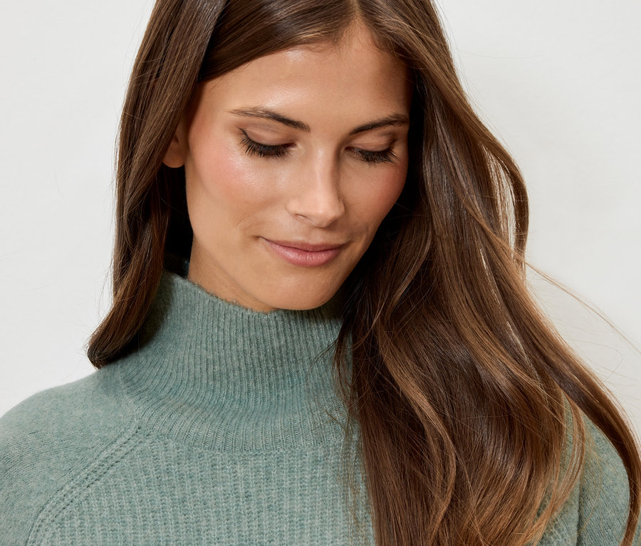 Kahverengi dalgalı saçlı, mint yeşili balıkçı yaka kazak giyen bir kadının portresi.