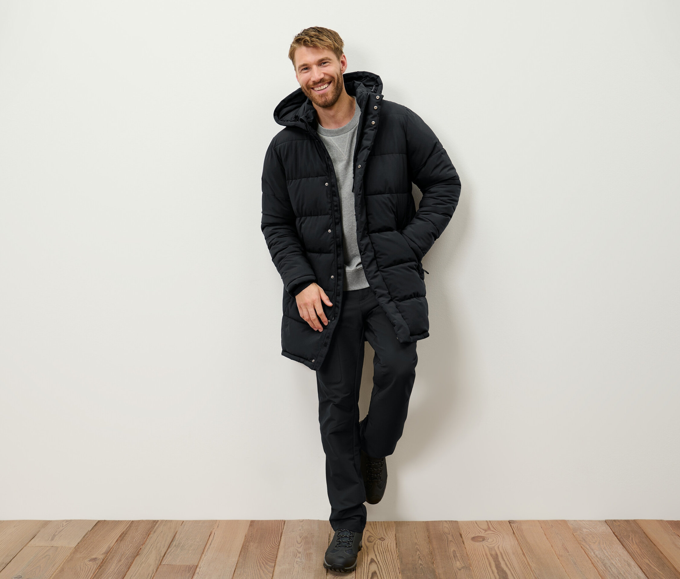 Gülümseyen sakallı bir adam duvara yaslanmış, siyah kapüşonlu şişme mont, siyah pantolon ve bot giyiyor.