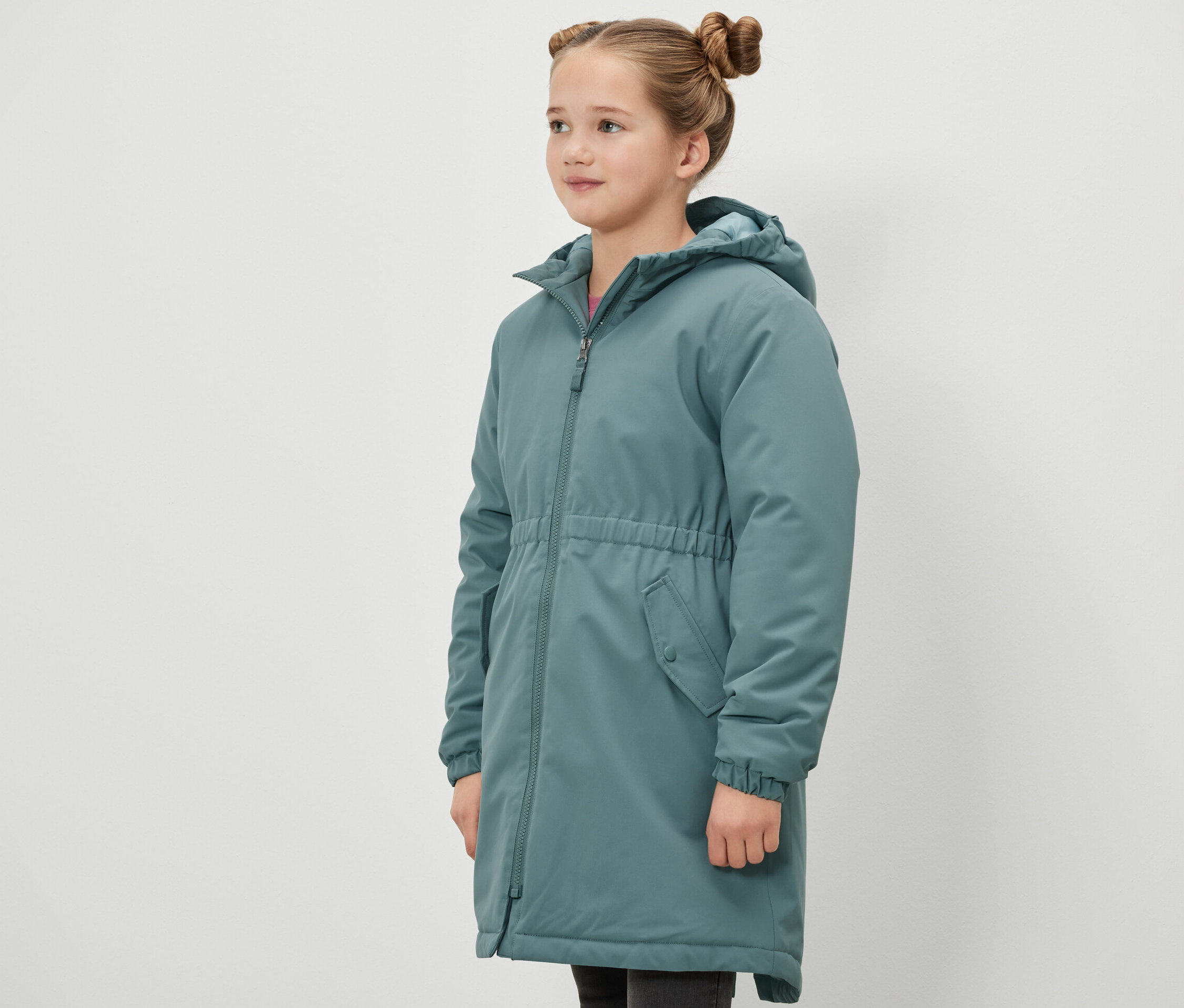 Kız çocuğu yan duruyor ve yeşil bir Kinder-Thermoparka termal parka giyiyor.