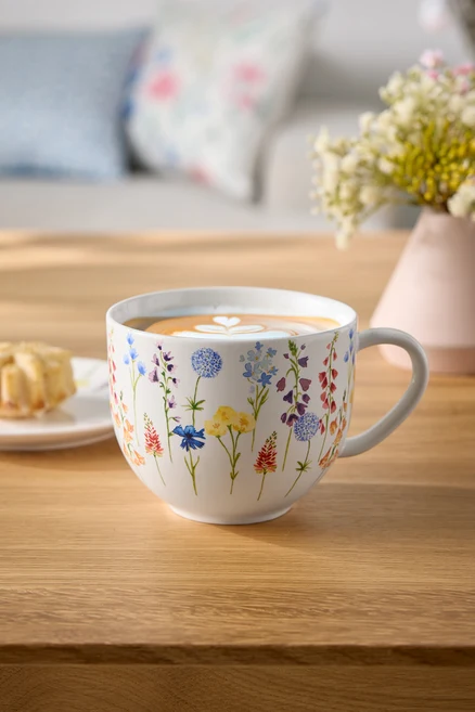 Çiçekli fincan, kahve ve latte sanatıyla ahşap bir masanın üzerinde, yanında bir tabak hamur işi ve çiçek vazosu bulunuyor.