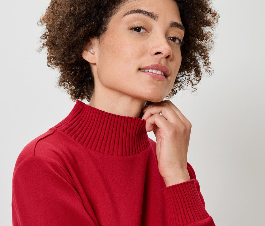 Kıvırcık saçlı, kırmızı balıkçı yaka kazak giyen ve eli boynunda olan bir kadının yakın plan portresi.