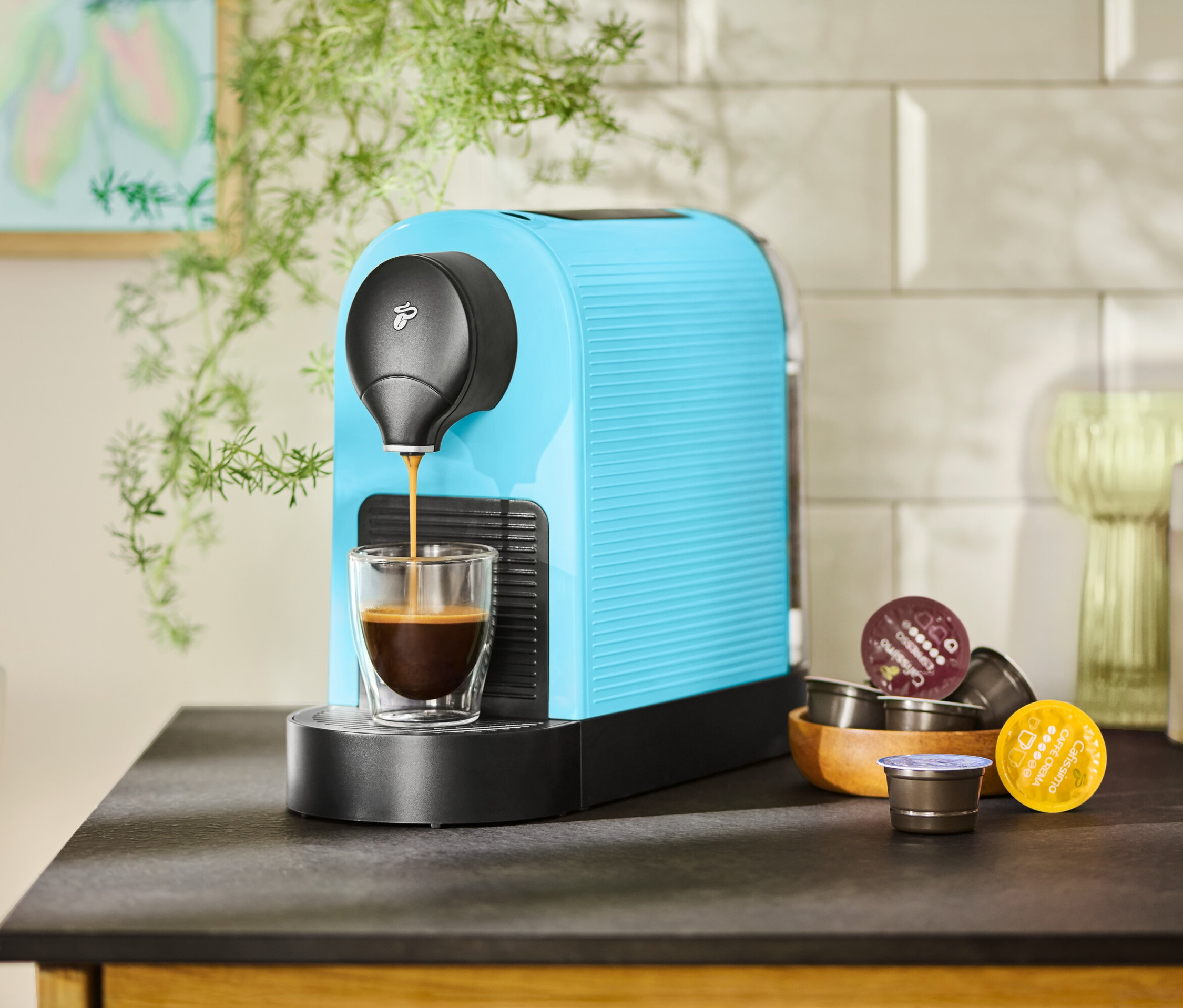 Turkuaz renkli bir kahve makinesi bir fincanı kahveyle dolduruyor. Yanında tahta bir kapta kahve kapsülleri duruyor.