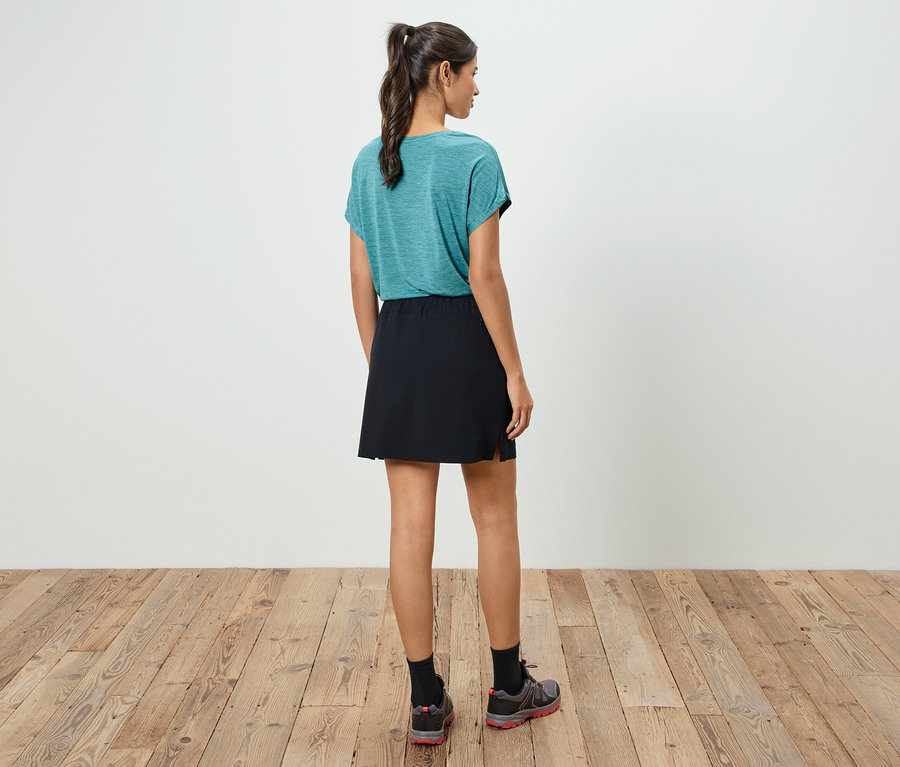 Bir kadın kameraya arkası dönük duruyor, üzerinde mavi-yeşil bir tişört, siyah bir etek ve spor ayakkabılar var.