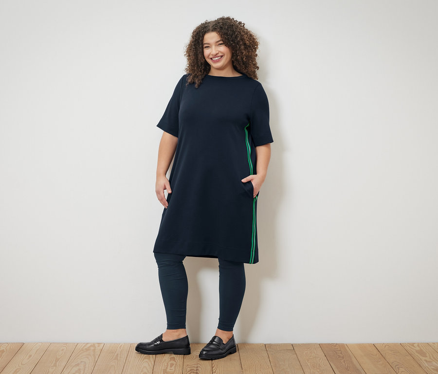 Kıvırcık saçlı bir kadın, üzerinde yeşil şeritli lacivert bir elbise, lacivert tayt ve siyah loafer ayakkabılar giyiyor.