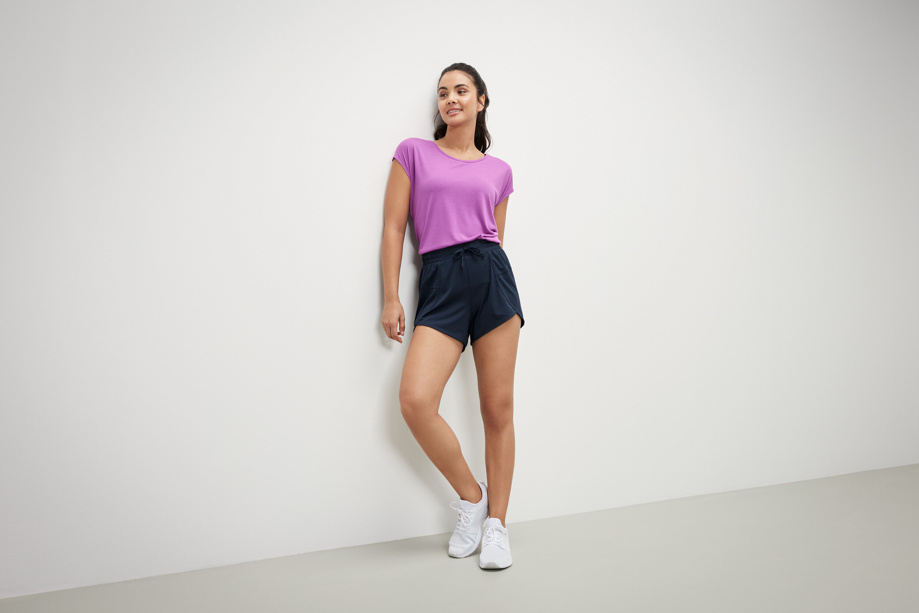 Duvara yaslanmış bir kadın mor tişört ve lacivert fonksiyonel şort giyiyor.