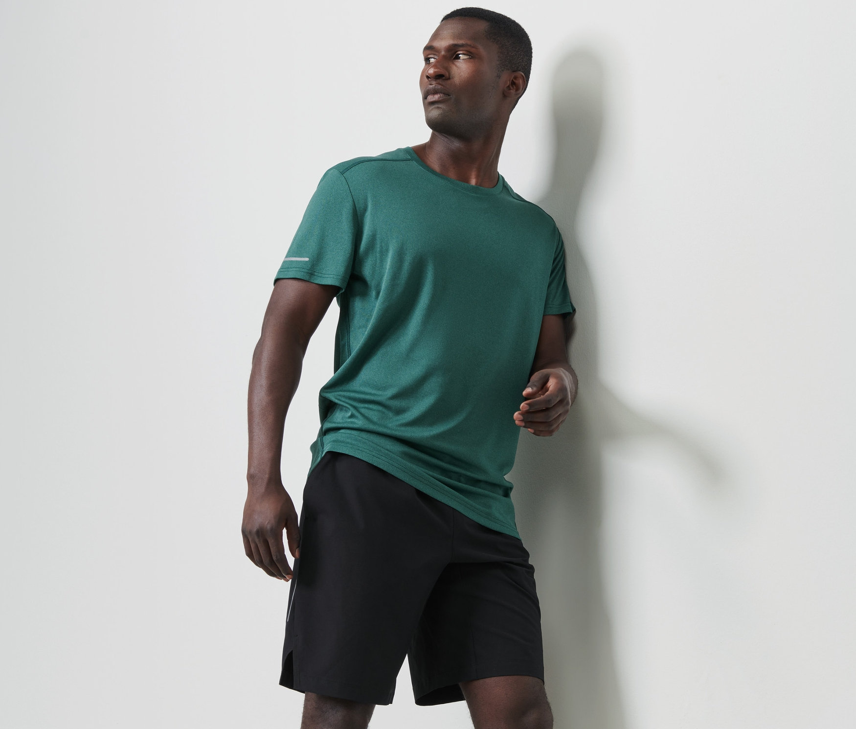 Duvara yaslanmış adam, kırçıllı koyu yeşil fonksiyonel tişört ve siyah şort giyiyor.