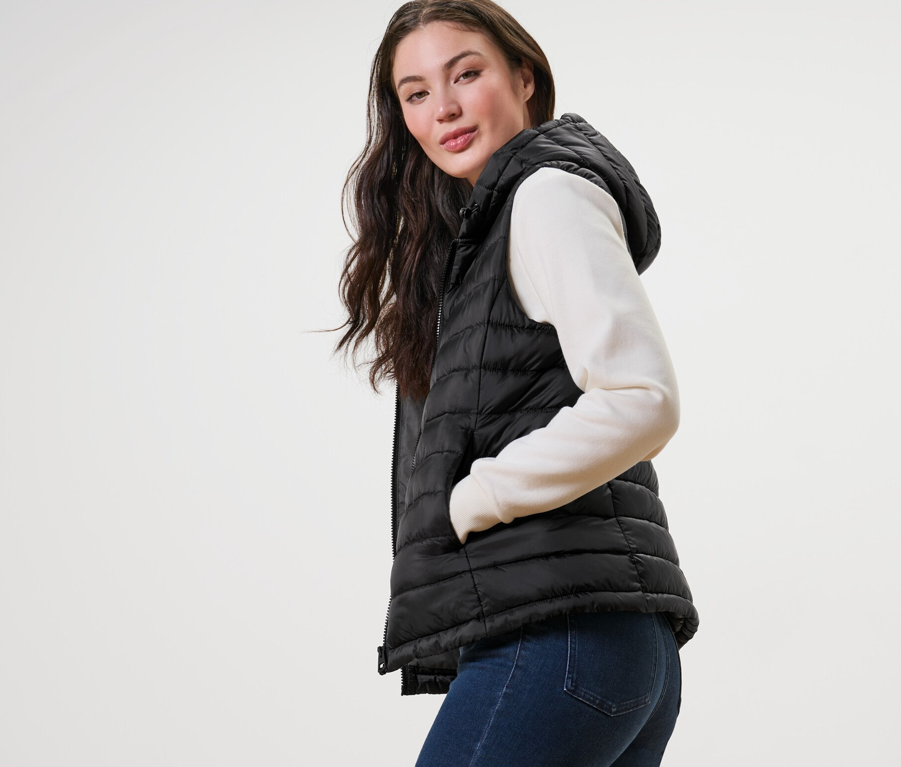 Kadın kapüşonlu kapitone yelek, kazak ve kot pantolonla poz veriyor.