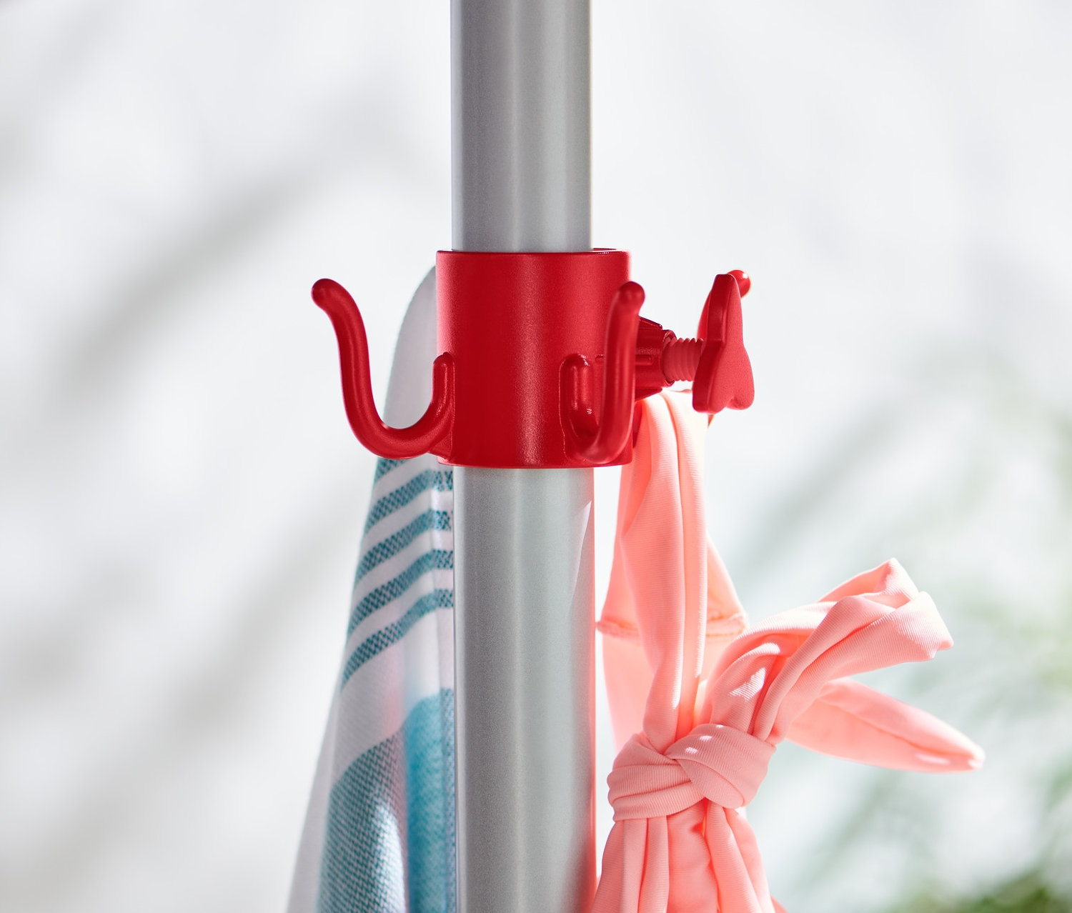 Gümüş renkli bir şemsiye direğine takılmış 2 adet kırmızı güneş şemsiyesi askısı üzerinde havlu ve pembe giysiler asılı duruyor.