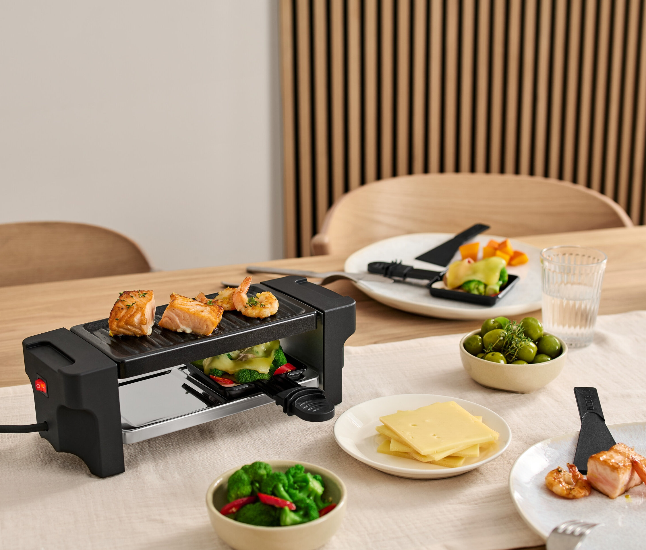 Masa üzerinde 2 Kişilik Raclette Izgara, ızgarada somon ve karidesler var. Malzemelerle dolu tabaklar ızgaranın etrafında duruyor.