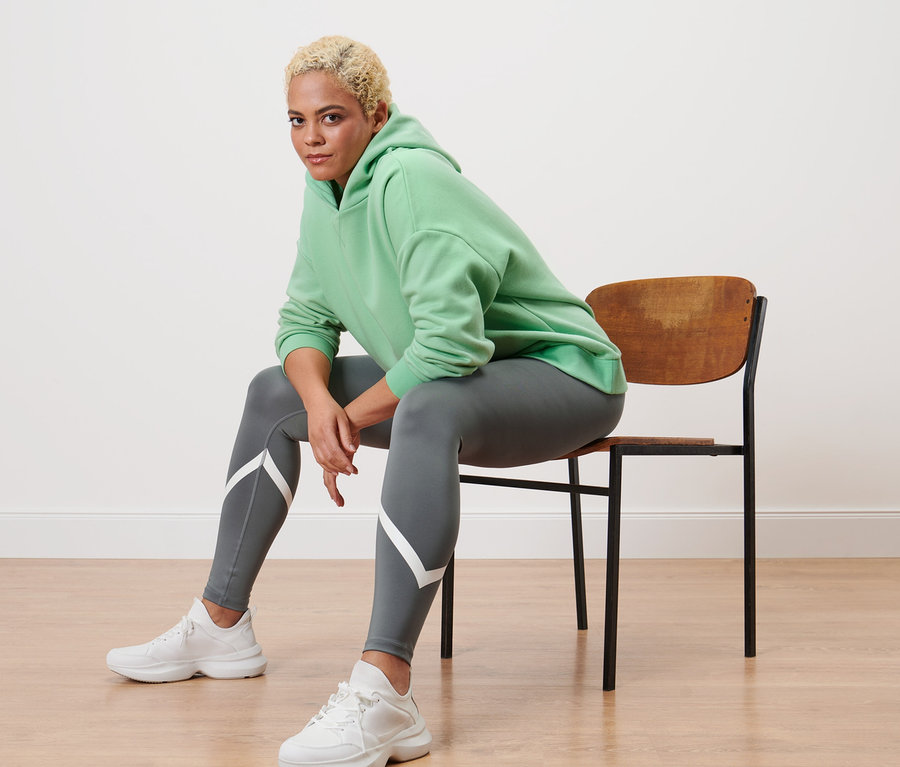 Bir kadın sandalyede oturuyor, açık yeşil bir sweatshirt ve gri spor taytı giyiyor.