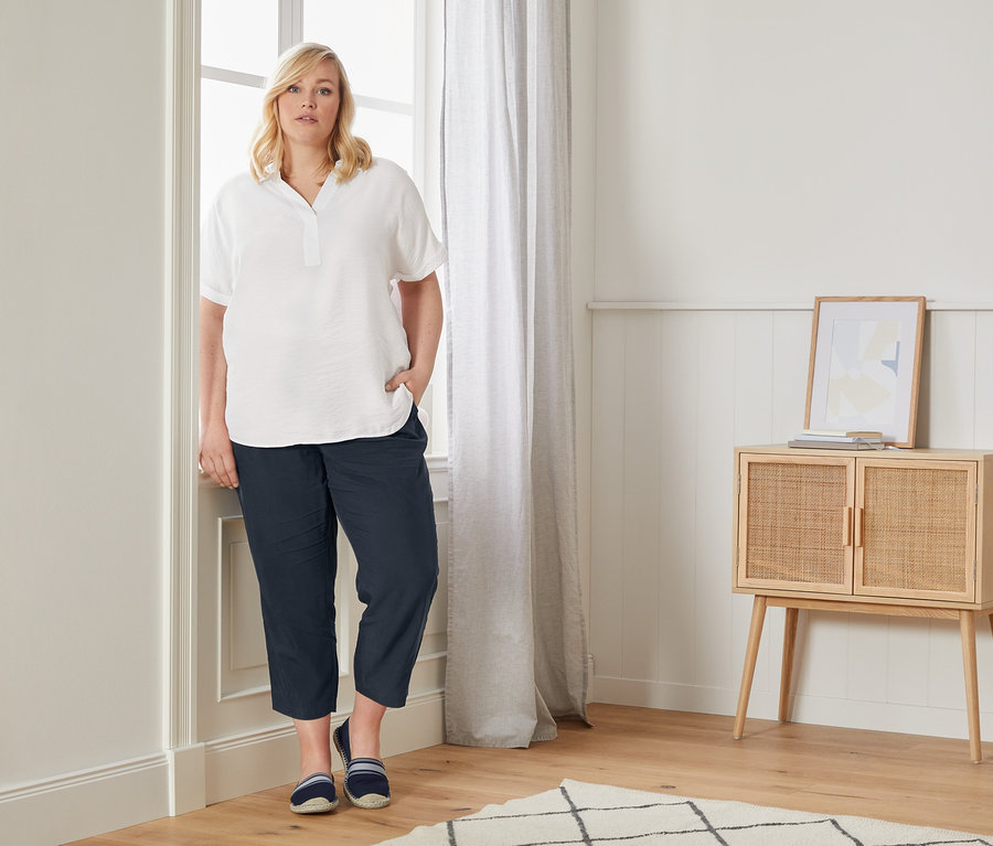 Bir kadın, pencere kenarında organik pamuklu oversized bir bluz, rahat chino pantolon ve espadril ayakkabılarla poz veriyor.