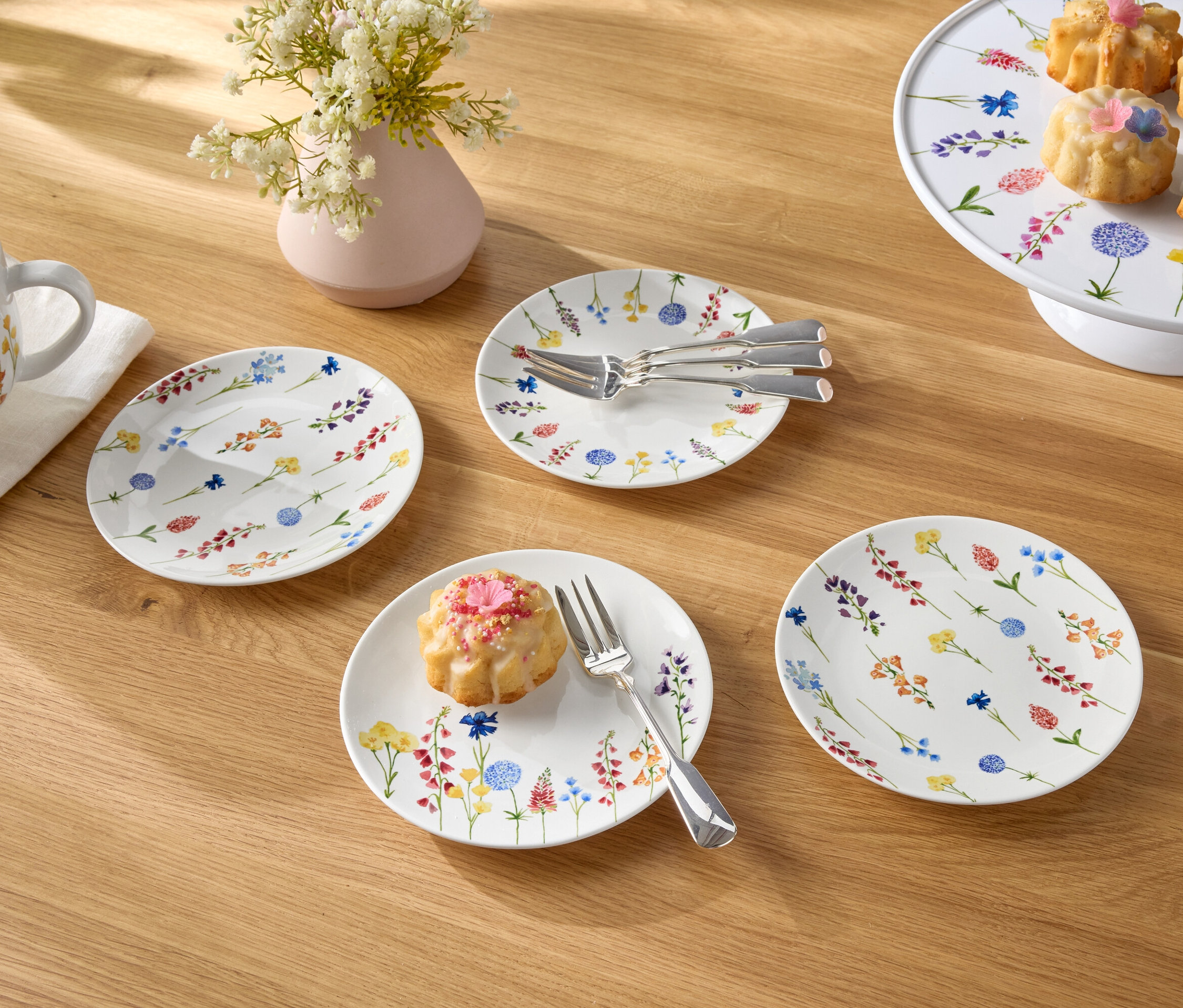 Ahşap bir masanın üzerinde bir fincan kahve, bir çiçek vazosu, tabaklar ve küçük kekler bulunmaktadır.