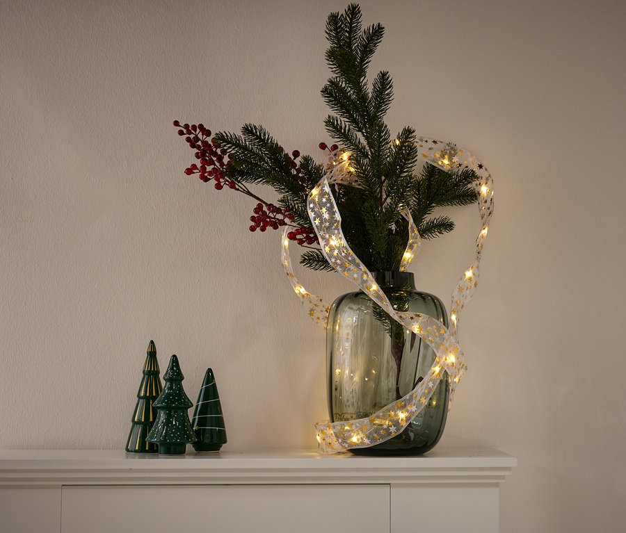 Vazoda çam ağacı dalları, kırmızı meyveler ve altın yıldızlı bir ışık zinciri var. Solda üç adet yeşil seramik ağaç duruyor.